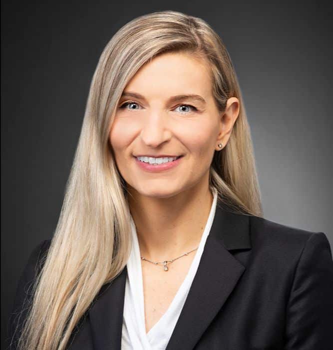 A woman with long, straight blonde hair is smiling and wearing a black blazer over a white blouse. She is posed against a dark, neutral background.
