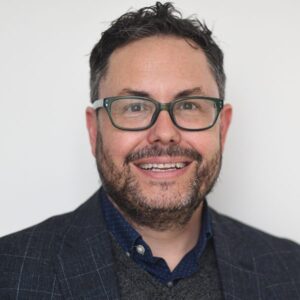 A person with short dark hair, glasses, and a beard smiles at the camera. They are wearing a dark blazer over a dotted shirt and sweater. The background is plain white.
